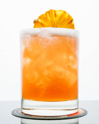 Closeup of orange cocktail with slice of dried pineapple out of focus, on the rim