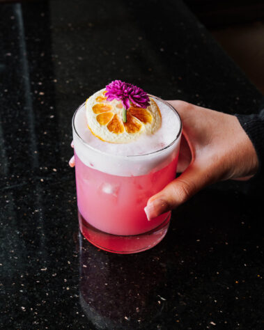 Hand holding a pink cocktail topped with an orange slice and purple flower