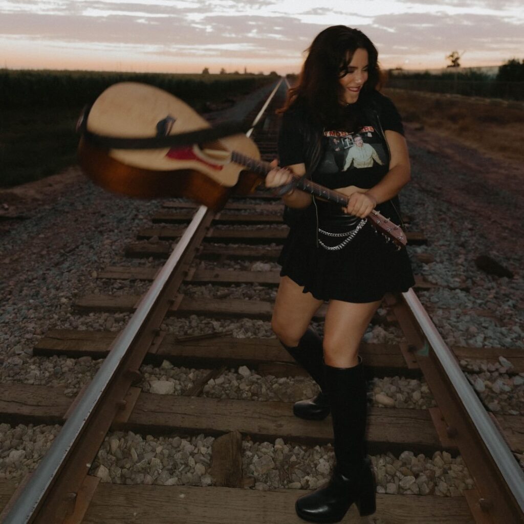 Sydnie Brazil holding an acoustic guitar over train tracks